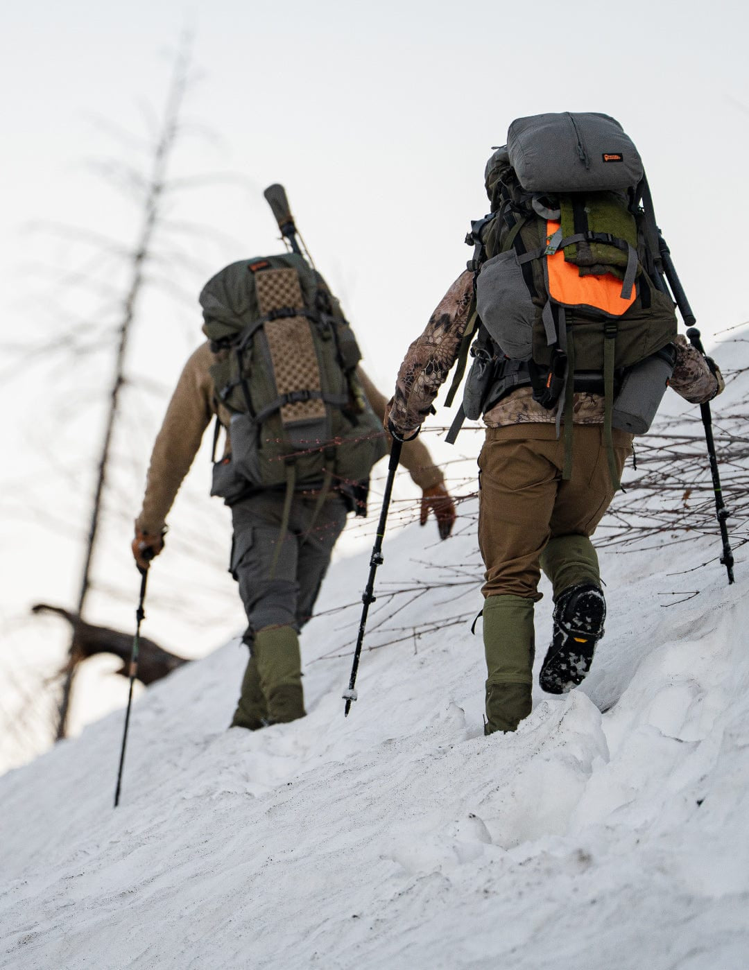 Two people hiking in the snow with backpacks with Backcountry Elite Trekking Poles and wearing Peax Storm Castle Hiking Gaiters.